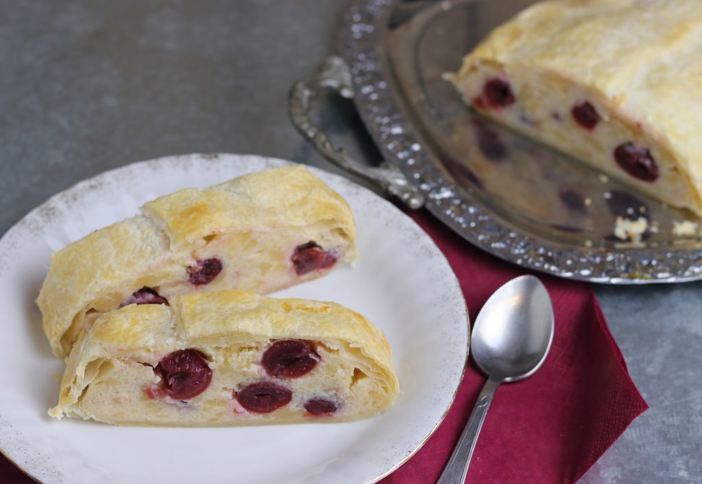 Quarkstrudel mit Kirschen. Der Strudel sieht zum Anbeißen aus und er ist absolut gelingsicher, da dieses Rezept mit fert. Zwei Stücke aufrecht auf einem Teller. Im Hintergrund der restliche Strudel.