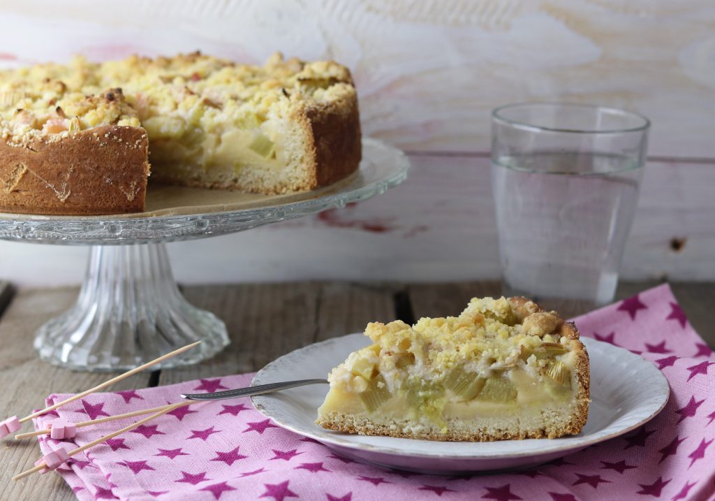 Rhabarberkuchen mit Streuseln und Puddingfüllung. Ein Stück auf einem Teller, dahinter der ganze Kuchen auf einer Etagere.