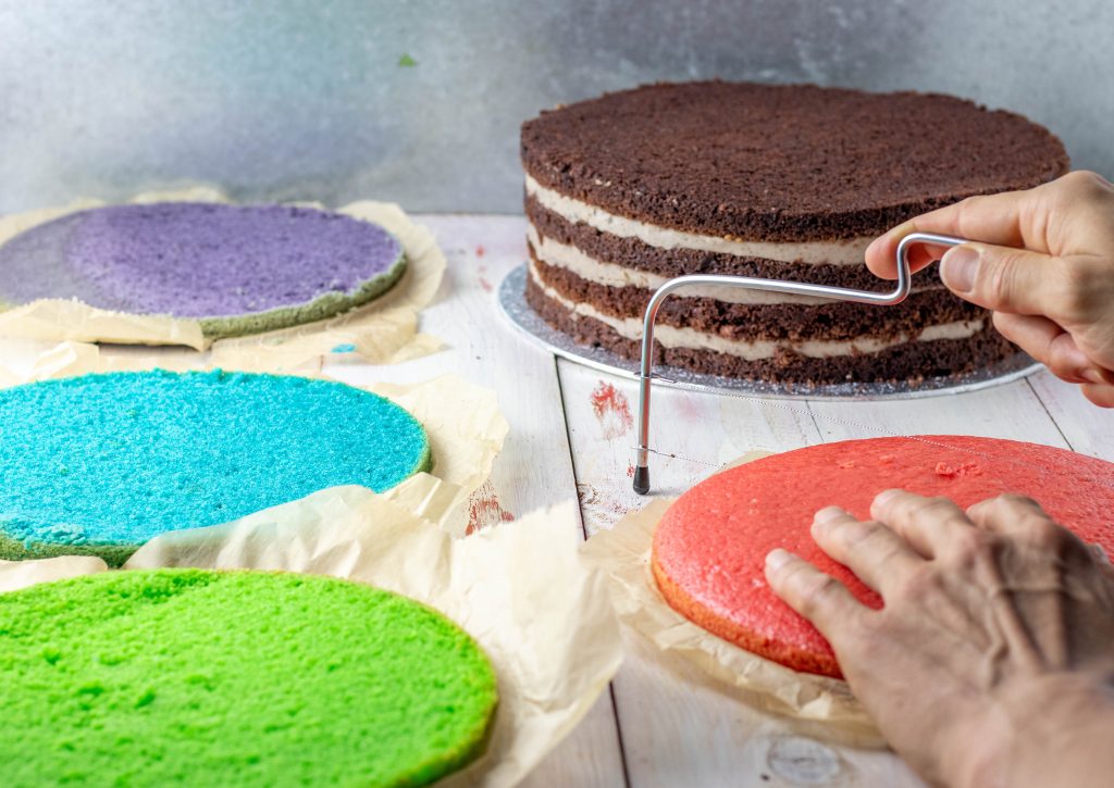 Um eine Torte zu schichten begradige ich die Biskuitböden. Im Hintergund sieht man eine geschichtete Torte mit Schokobiskuit. Im Vordergrund schneide ich den Regenbogenbsikuit. Der Regenbogenkuchen wird die zweite Torte einer dreistöckigen Hochzeitstorte.