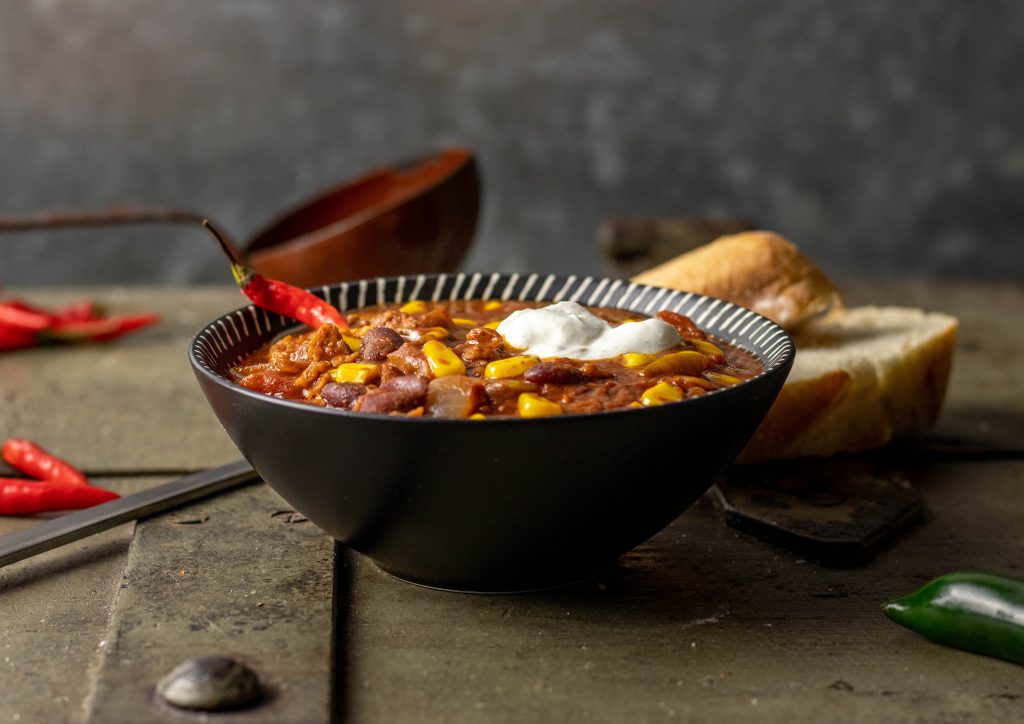 Chili sin Carne in einer Schüssel, auf einer alten Truhe mit einer Suppenkelle, Brot und Chilischoten