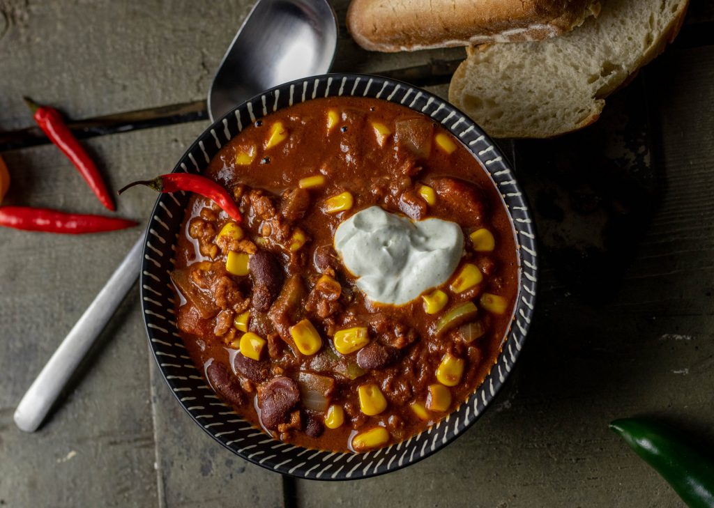 Chili sin Carne in einer Schale, daneben Brot, Chilis und ein Löffel
