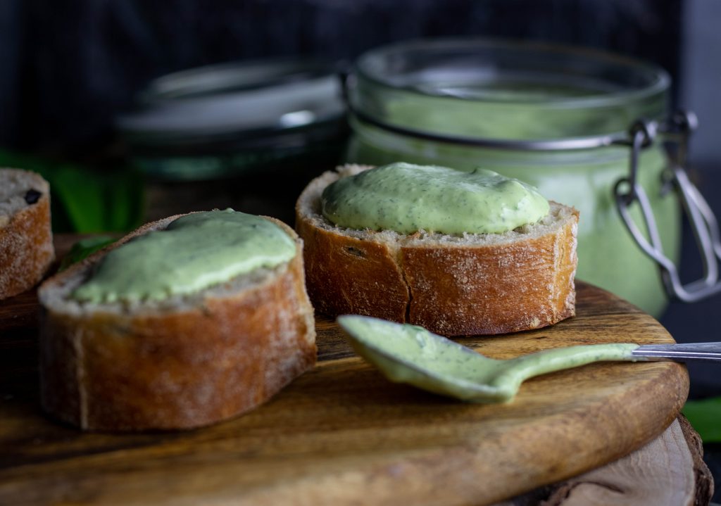 Bärlauch Aioli auf Baguette. Außerdem ein Glas und ein Löffel mit Bärlauch Aioli.