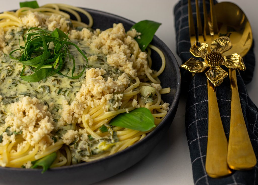 Cremige Limetten Bärlauch Sauce mit veganem Parmesan auf einem Teller. Daneben goldenes Besteck mit Serviettenring.