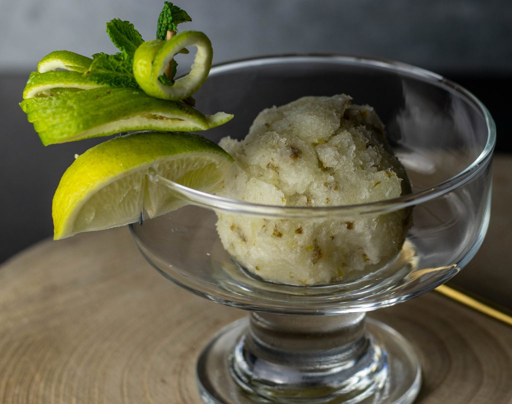 Eine Kugel Limetten-Minz-Sorbet in einem Glasbecher. Mit frischem Minz- und Limettendekor am Rand.