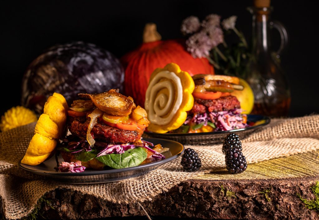 Reichhaltig belegte Herbstburger, dahinter herbstliches Gemüse, Blumen und Öl.