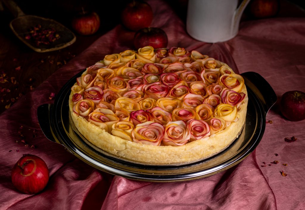 Apfelrosentarte auf einem Kuchenteller auf einer rosa Tischdecke.