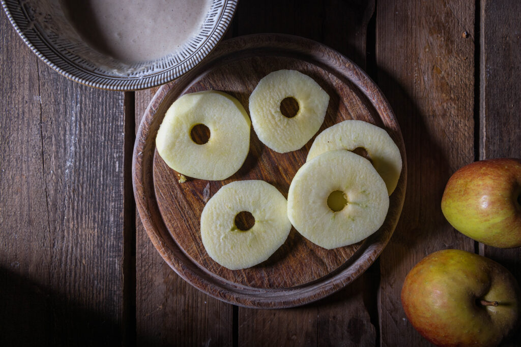 Geschälte Apfelscheiben ohne Kerngehäuse, daneben 2 Äpfel und eine Schüssel mit Teig.