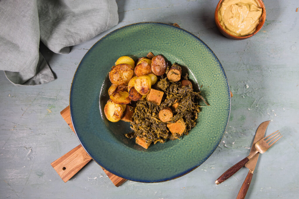 Veganer Grünkohl mit selbstgemachtem, pflanzlichen Kassler und Würstchen auf einem grünen Teller.