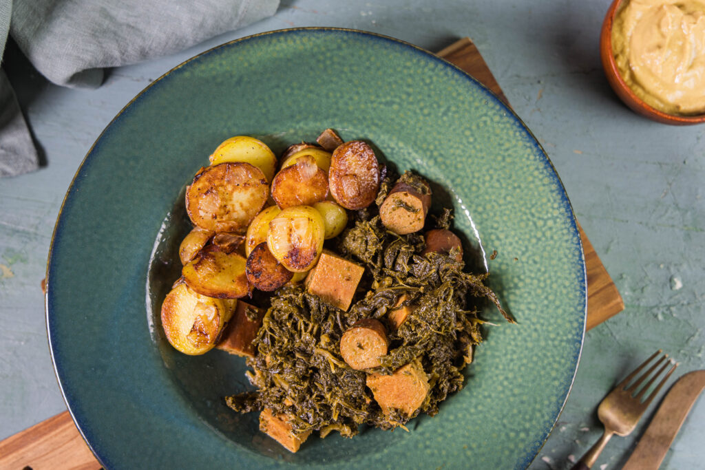 Veganer Grünkohl mit selbstgemachtem, pflanzlichen Kassler und Würstchen auf einem grünen Teller.