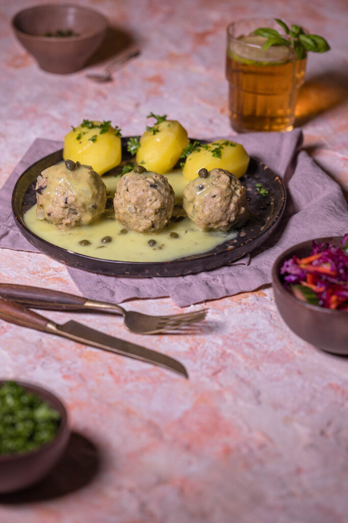 Ein rosraroter Tisch mit einem Trinkglas, mehreren Schalen, Besteck und einem Teller mit Königsberger Klopsen, Kartoffeln und Klopssauce.