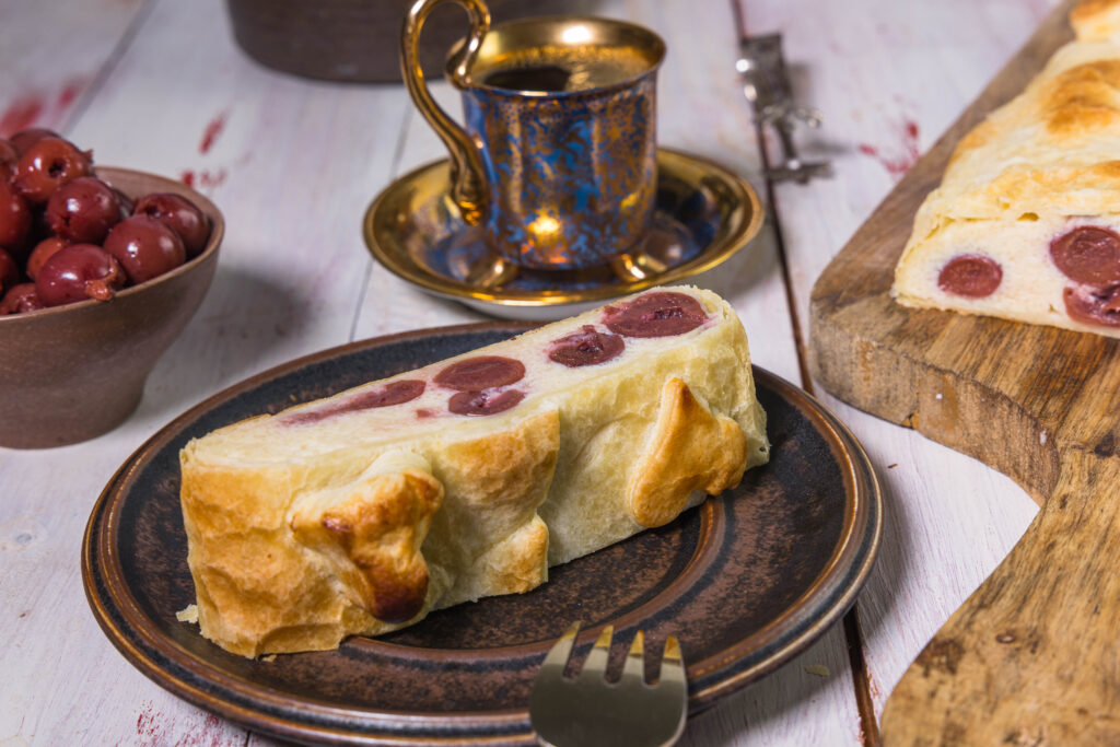 Ein Stück veganer Quarkstrudel mit Kirschen auf einem Teller. Der Teig ist mit Blätterteigherzen verziert. Dahinter eine Sammeltasse mit Esspresso.