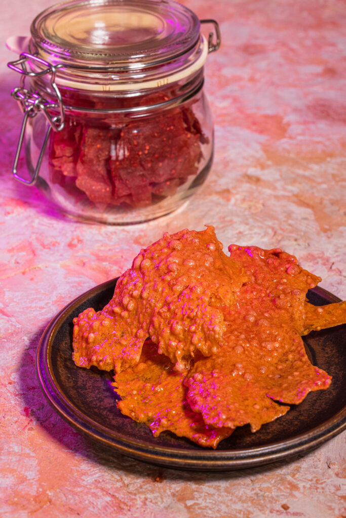 Gepuffte Rote Beete Cracker auf einem braunen Teller. Dahinter ein Glas mit unfrittierten Crackern.