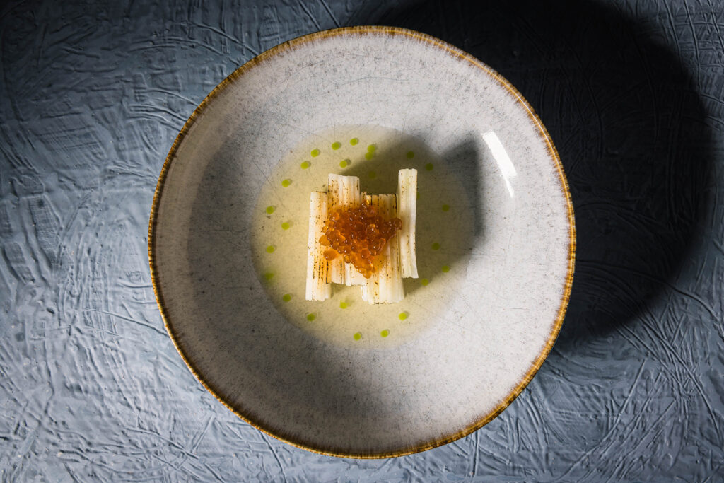 Kohlrabisalat in einem tiefen Teller. Der Kohlrabi ist gehobelt und als in sich versetzter Quadar angeordnet. Getoppt von Zwiebel-Kaviar. Um den Kohlrabi ist Holunderessig mit Tropfen aus Schnittlauchöl.