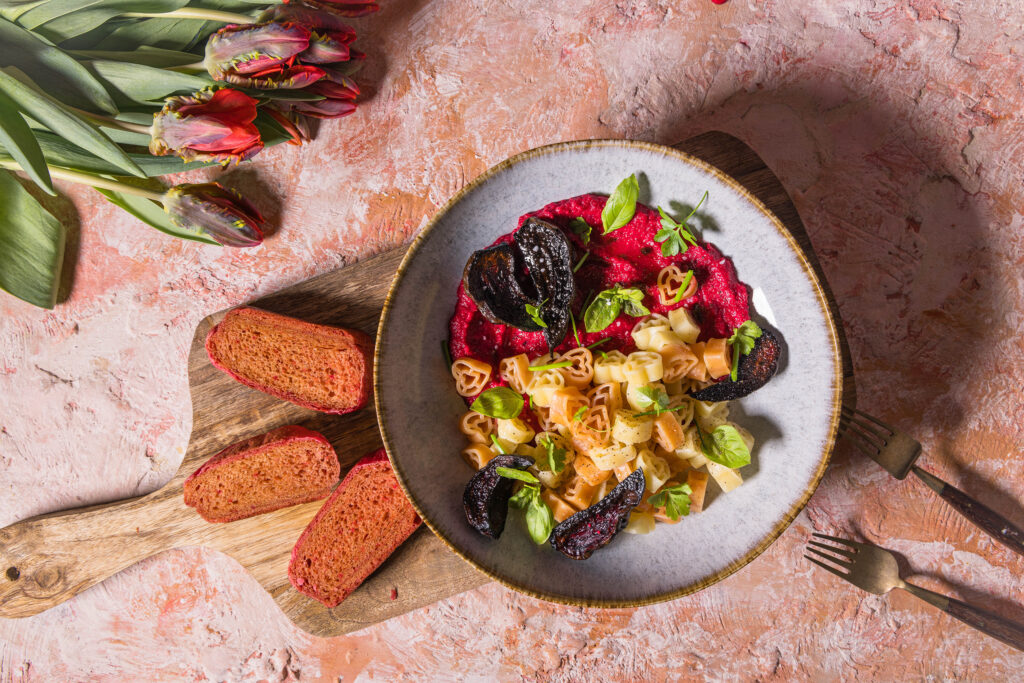 Herzchenförmige Pasta mit Rote Beete Hummus Sauce. Daneben drei Scheiben rosa Pfannenbrot und Tulpen.