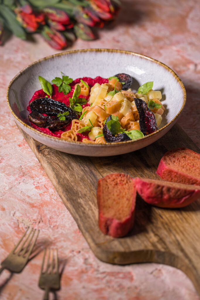 Herzchenförmige Pasta mit Rote Beete Hummus Sauce. Daneben drei Scheiben rosa Pfannenbrot und Tulpen.