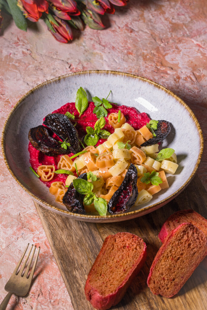Herzchenförmige Pasta mit Rote Beete Hummus Sauce. Daneben drei Scheiben rosa Pfannenbrot und Tulpen.