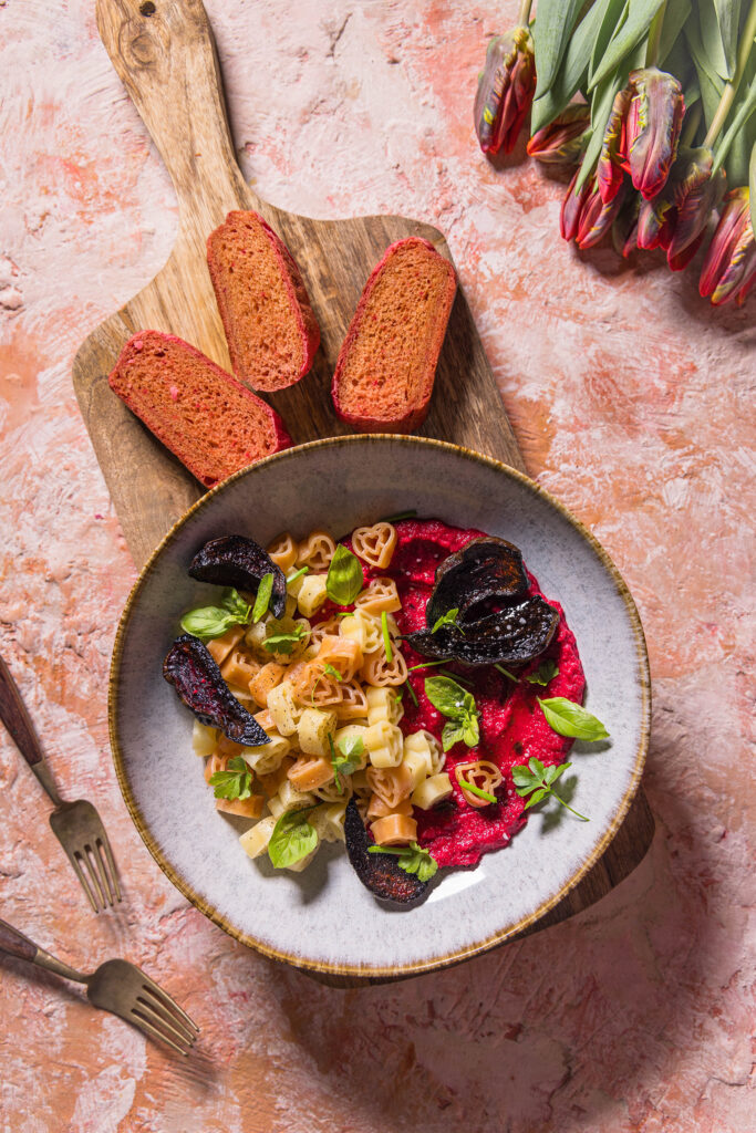 Herzchenförmige Pasta mit Rote Beete Hummus Sauce. Daneben drei Scheiben rosa Pfannenbrot und Tulpen.