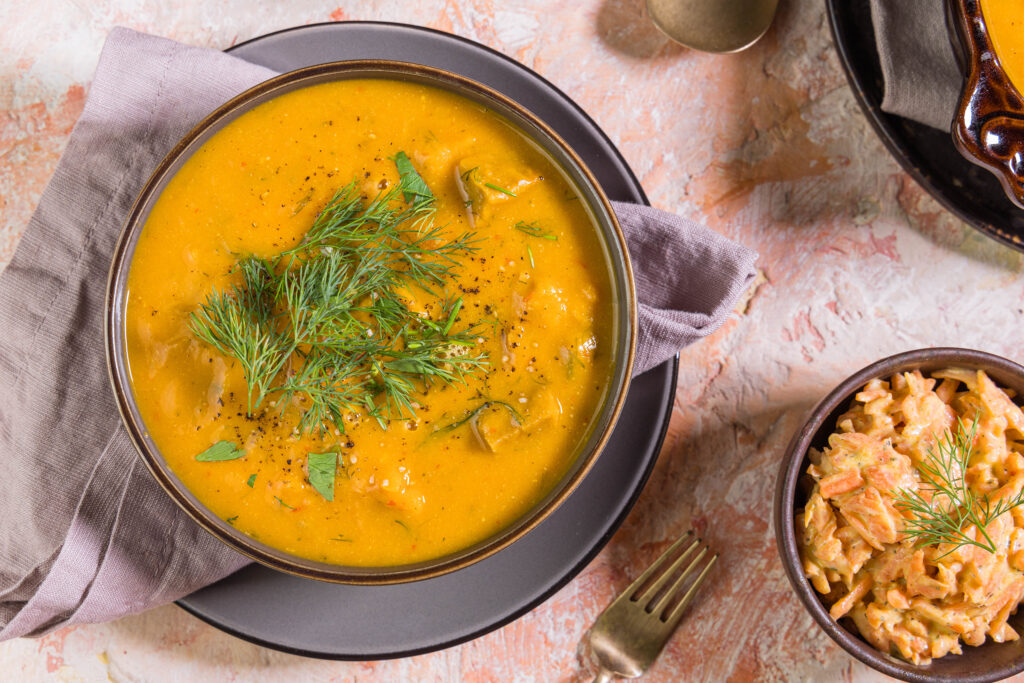 Süßkartoffel-Kokos-Suppe in einer Schüssel, mit Dill garniert, daneben ein Karottensalat