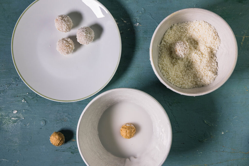 Eine Schale mit Zuckerguss und einer Waffelkugel darin, eine Schale mit Kokosflocken und einem Vaffaello darin und ein Teller mit drei fertigen veganen Raffaellos.