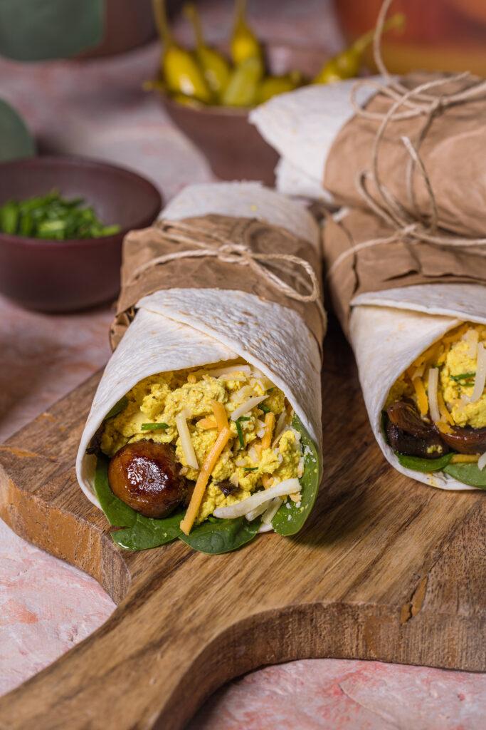 Breakfast Burritos auf einem Holzbrett, dahinter Schalen mit Peperonis und Kräutern.