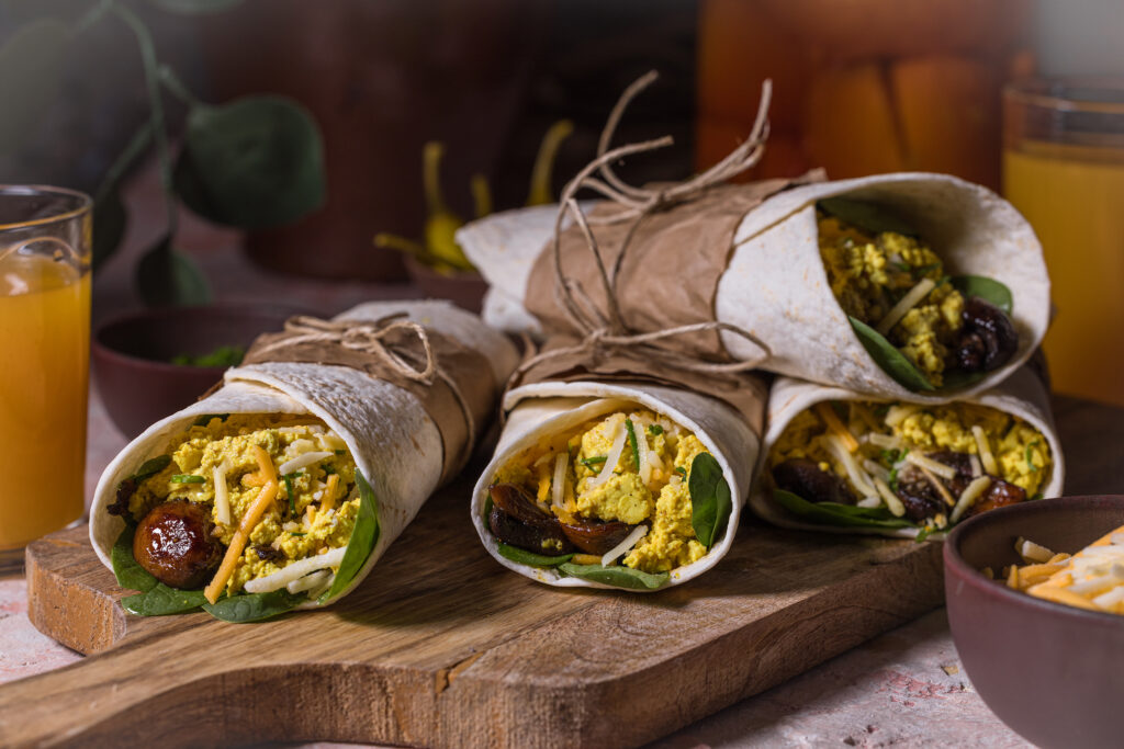 Vegane Egg and Cheese Breakfast Burritos auf einem Holzbrett, Gläser mit Saft und eine Schale veganer Reibekäse.