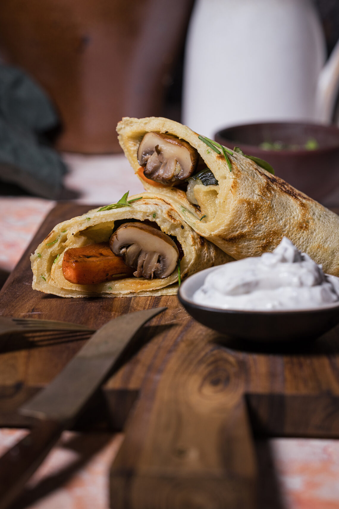 Gefüllte Kräuter-Pfannkuchen, vegan, mit Grillgemüse und Dip auf einem Holzbrett. Davor Besteck.