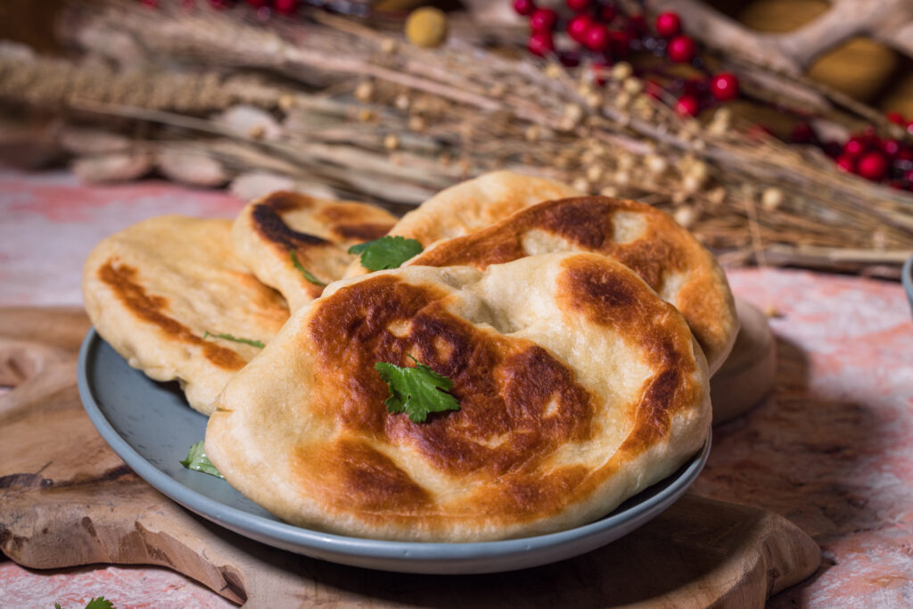 Mehrere Naan auf einem Teller. Garniert mit Koriander. Im Hintergrund einige Trockenblumen.