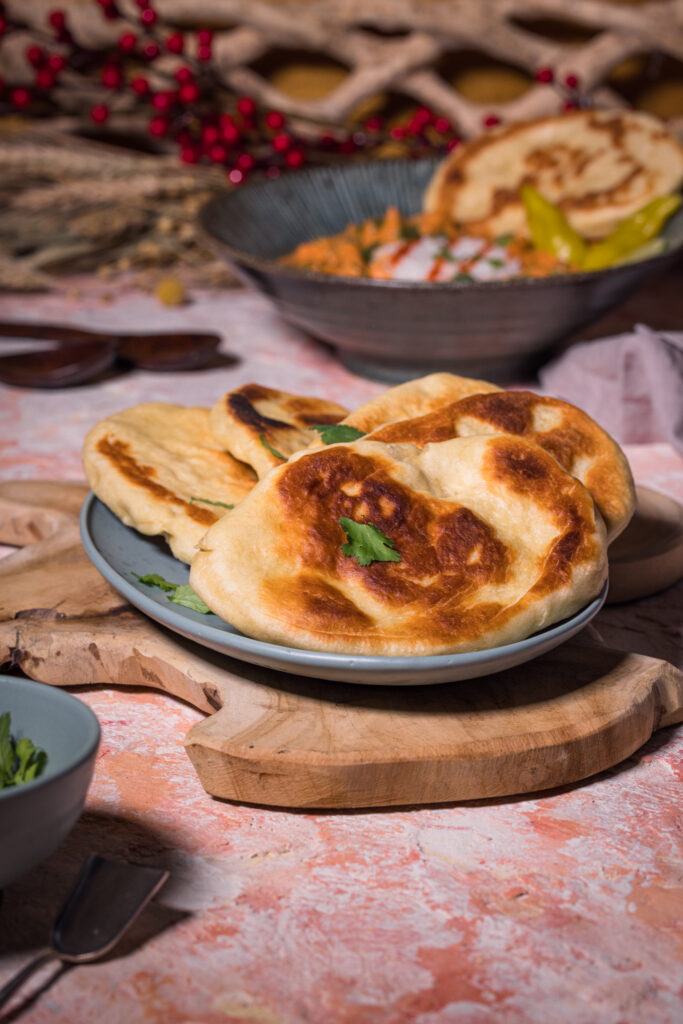 Mehrere Naan auf einem Teller. Garniert mit Koriander. Im Hintergrund einige Trockenblumen und ein Teller mit Seitan Tikka Masala.