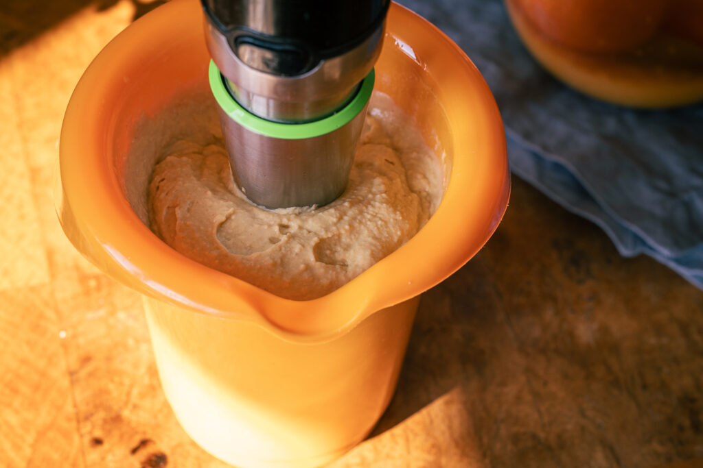 Ein Pürierstab püriert einen cremigen Hummus in einem hohen Gefäß