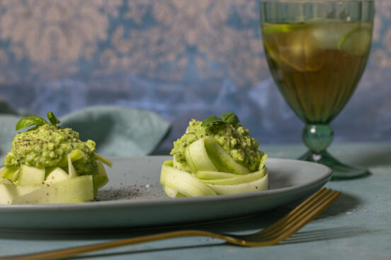 Zucchini-Tagliatelle mit Avocado-Pesto auf einem grünen Teller, ein Getränk mit Limetten im Hintergrund