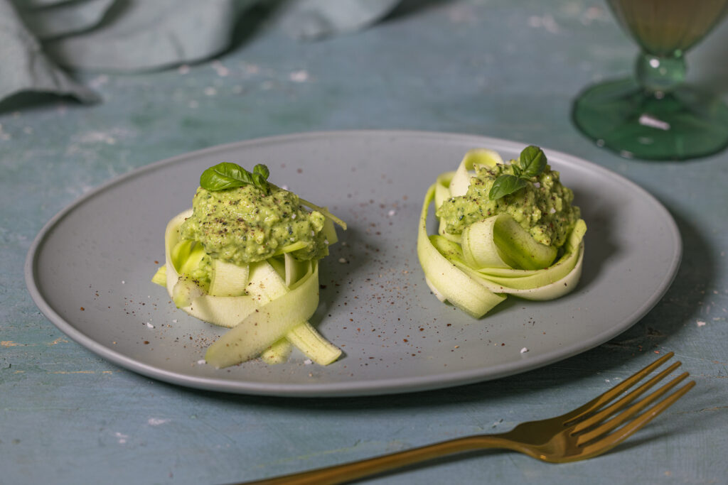 Zucchini-Tagliatelle mit Avocado-Pesto auf einem grünen Teller, ein Getränk mit Limetten im Hintergrund
