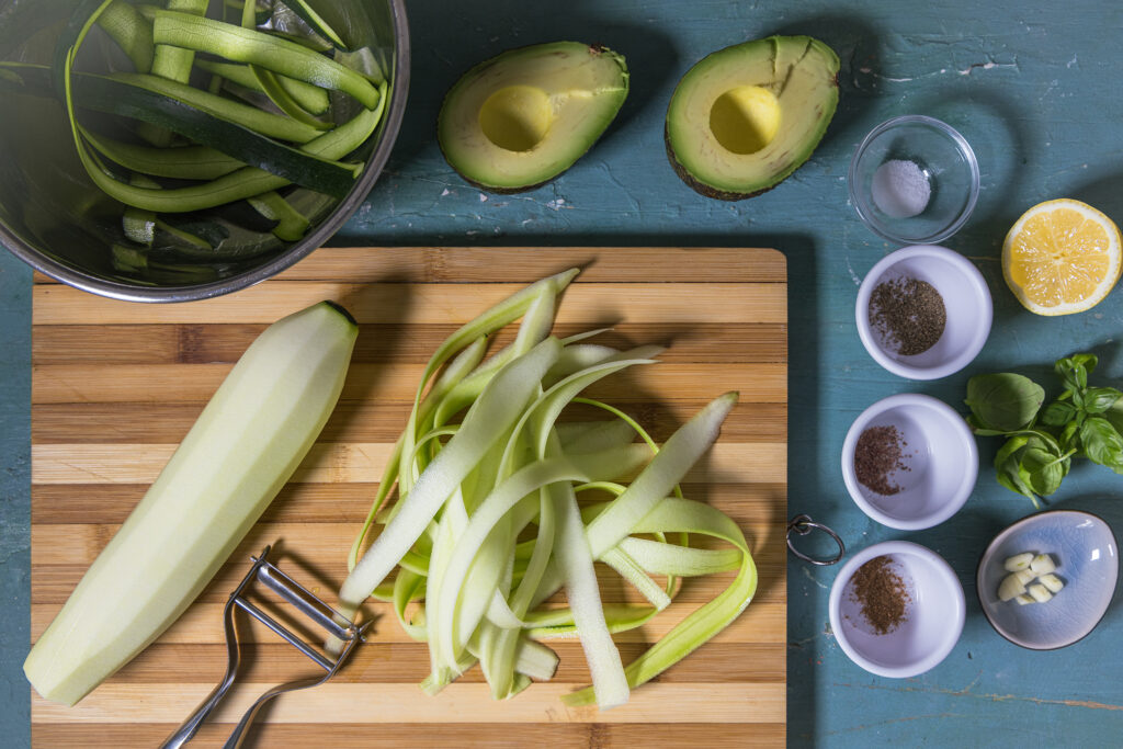 Eine Zucchini wird in Streifen geschält. Um das Brett herum sind weitere Zutaten und eine Schale mit der Zucchinischale.