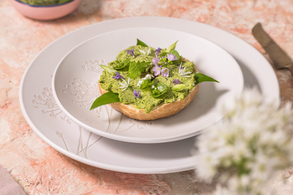 Bärlauchstreich auf einem krossen Bagel - zart garniert mit Bärlauchblüten, Knoblauchsruke, Wiesenschaumkrautblüten, Tellerkraut, Bärlauchknospen und Grundermannblüten