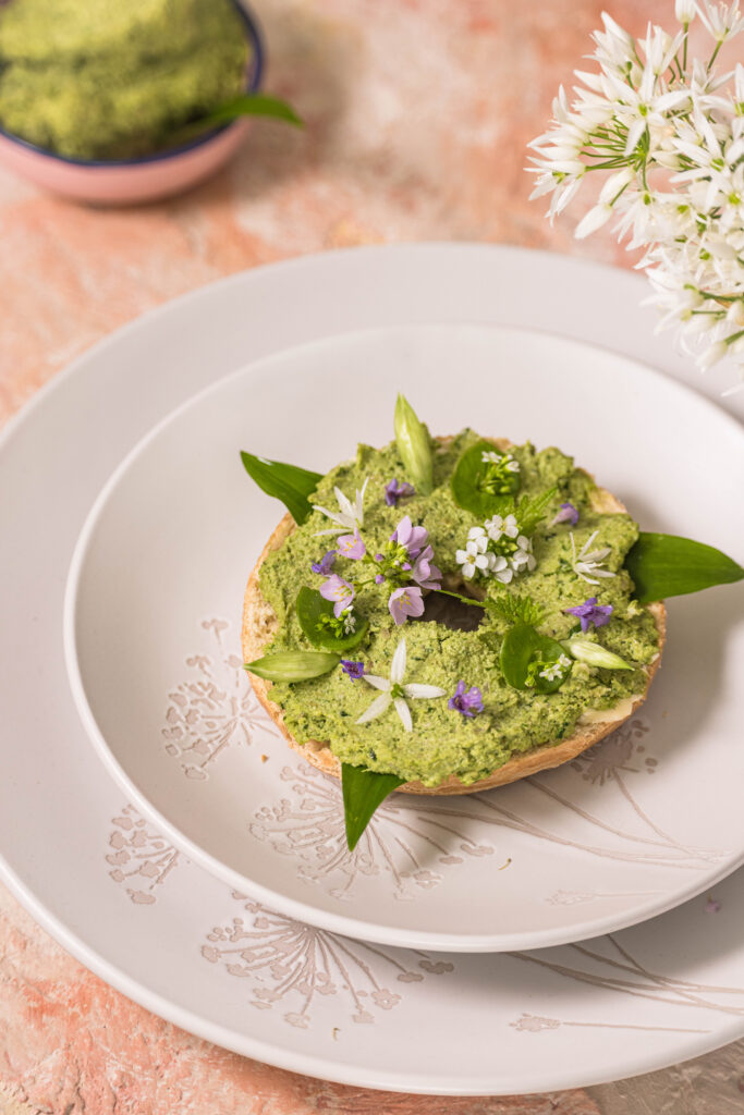 Bärlauchaufstrich auf einem krossen Bagel - zart garniert mit Bärlauchblüten, Knoblauchsruke, Wiesenschaumkrautblüten, Tellerkraut, Bärlauchknospen und Grundermannblüten