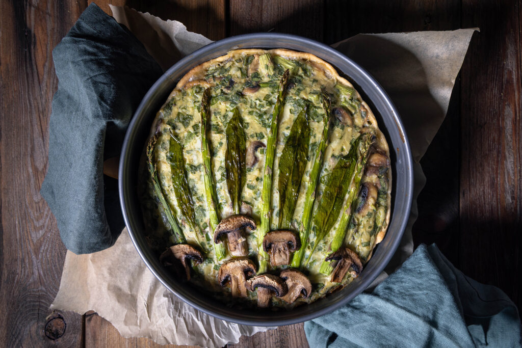 Eine frisch gebackene vegane Bärlauchquiche mit grünem Spargel und Champignons in einer Backform.