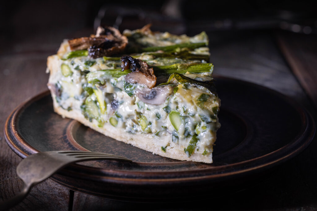 Bärlauchquich mit Spargel, Bärlauch und Champignons, ein Stück auf einem braunen Teller.