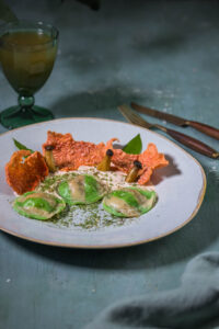 Bärlauch-Ravioli auf hellem Tomatenschaum mit Rote Beete Crackern und kleinen Kräutersetilingen auf einem Teller.