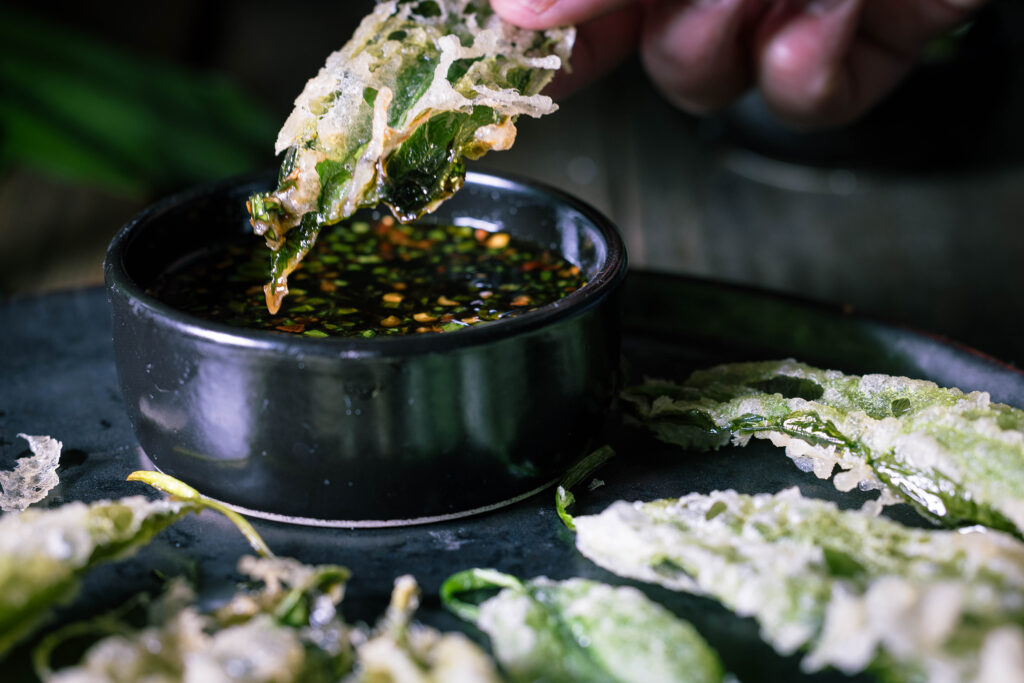 Ein in Tempura-Teig frittiertes Bärlauchblatt wird in einen Dip getunkt.