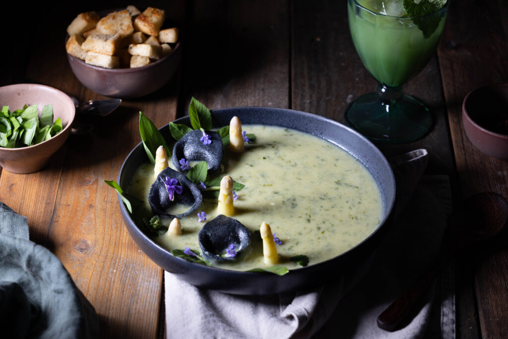 Bärlauch-Kartoffel-Cremesuppe mit Bärlauch-Tortellini und Spargel. Daneben eine Schale Croutons und frischer Bärlauch.