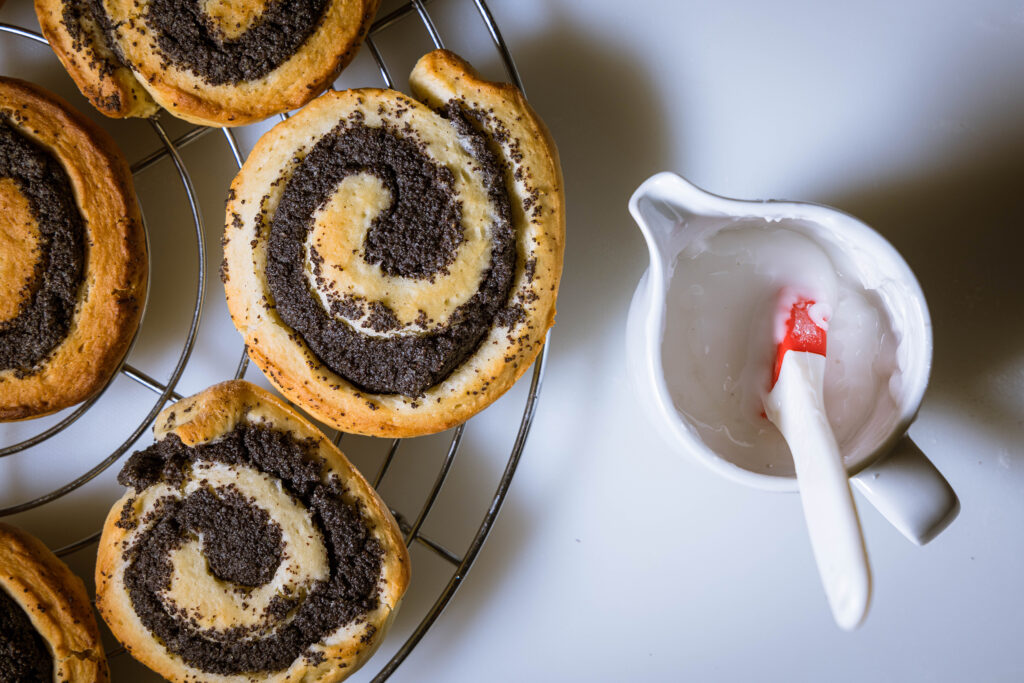 Mohnschnecken kühlen auf einem Kuchengitter aus. Daneben ein Kännchen mit Zuckerguss.