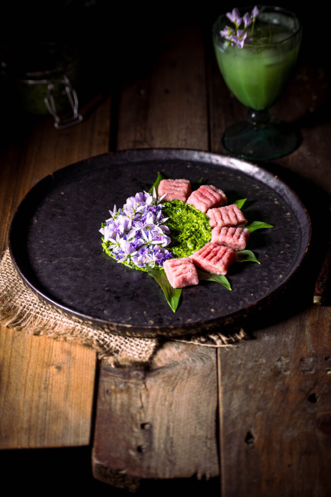 Rote Beete Gnocchi mit Bärlauchpesto und essbaren Blüten von Wildkräutern angerichtet auf einem dunklen Teller.