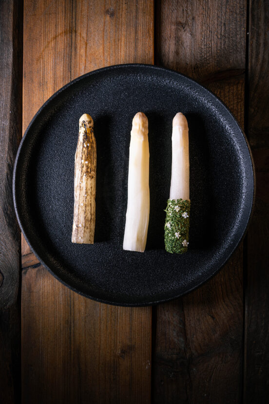 Dreierlei Spargel – fermentiert, mit Holunderblüte und flambiert - angerichtet auf einem dunklen Teller