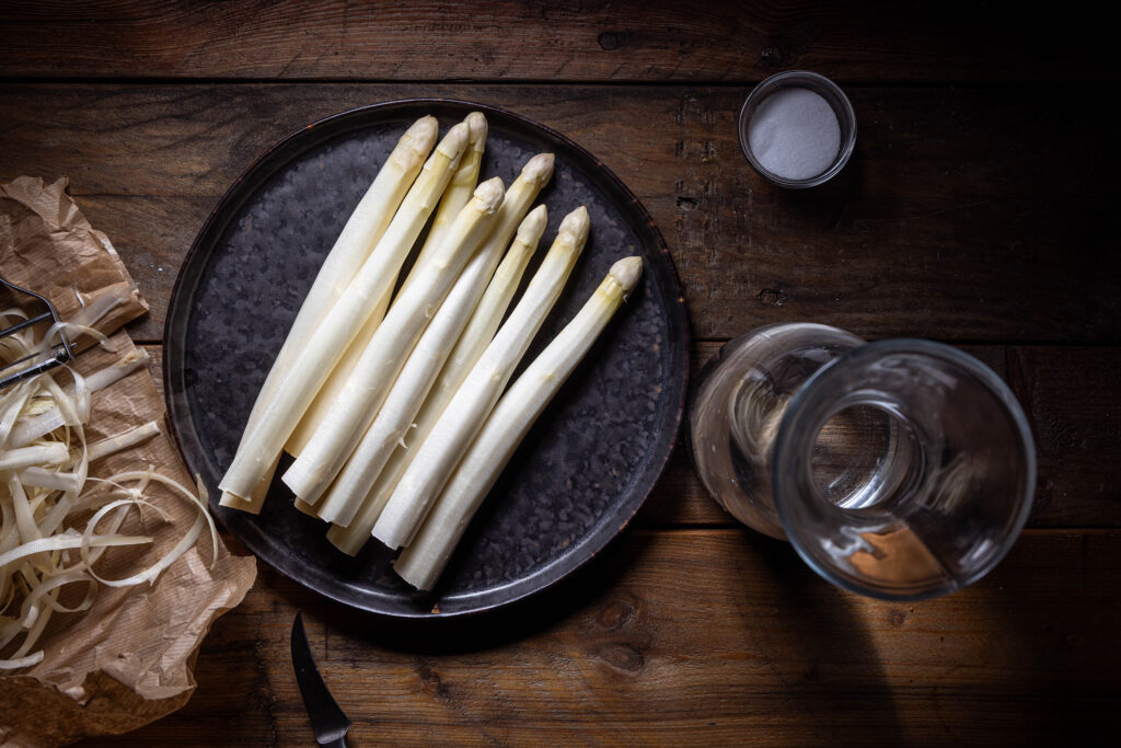 Geschälter Spargel auf einem dunklen Teller. Daneben eine Karaffe mit Wasser, eine Schale mit Salz und die Spargelschalen auf einem braunen Papier.