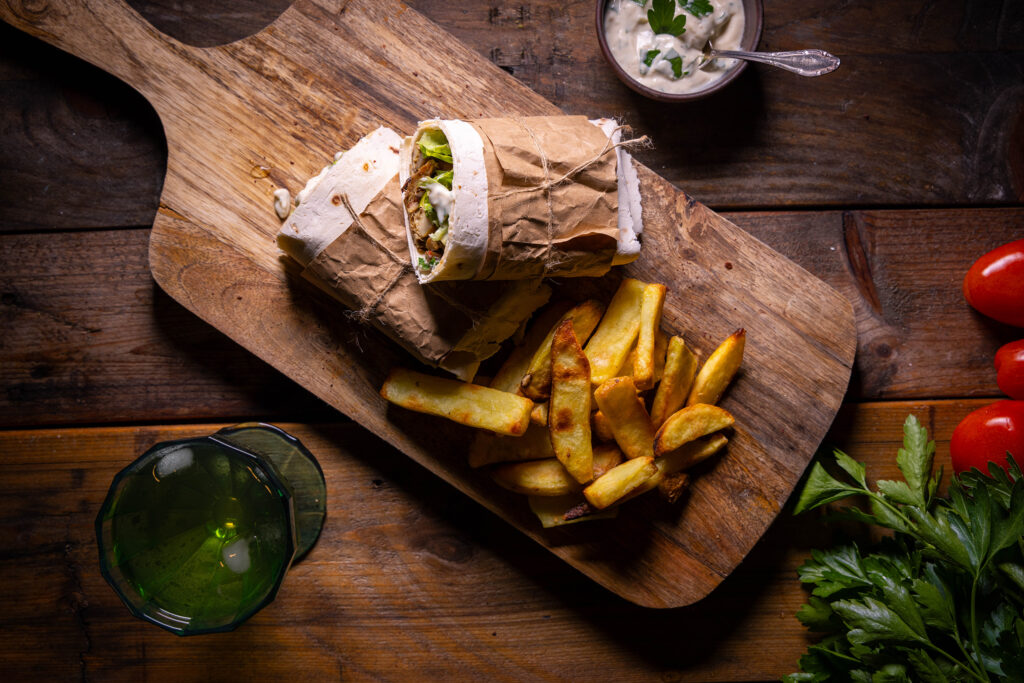 Vegane Schawarma mit Grillgemüse und Joghurt-Tahini-Sauce mit Pommes auf einem Holzbrett. Daneben eine Schale Joghurt-Tahini-Sauce, ein Glas und Gemüse.
