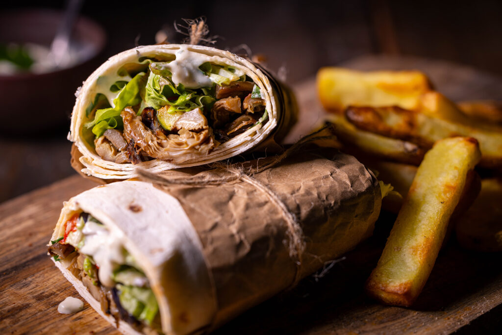 Vegane Schawarma aus Austernseitlingen mit Grillgemüse. In Papier gewickelt auf einem Holzbrett mit knusprigen Pommes als Beilage.