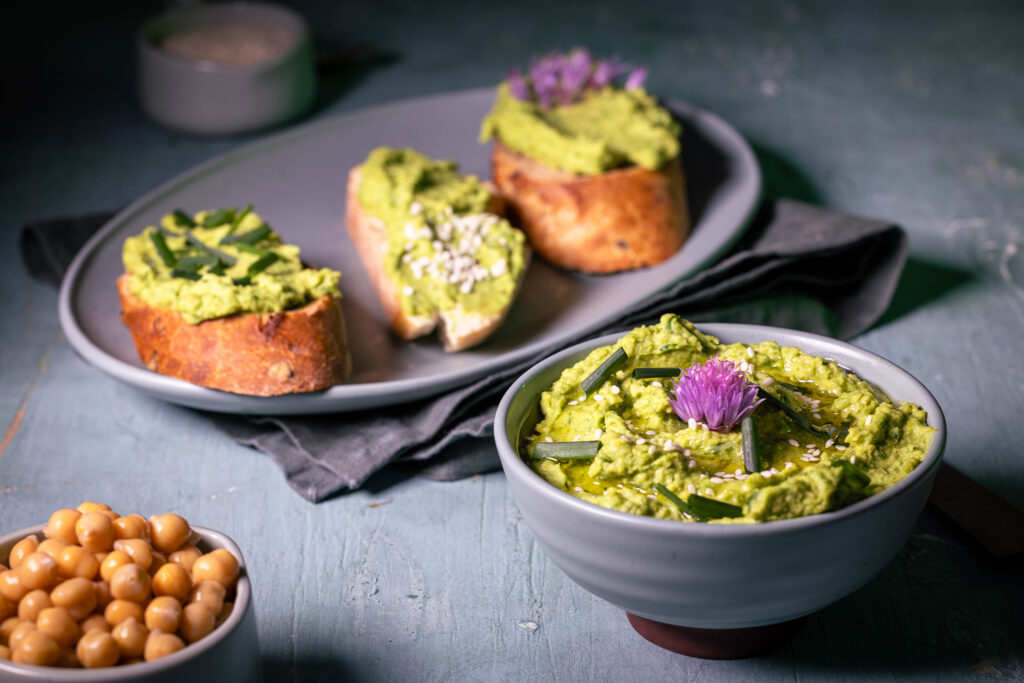 Grüner Hummus mit frischem Schnittlauch in einer Schale und auf Brot. Außerdem ein Schälchen Kichererbsen.