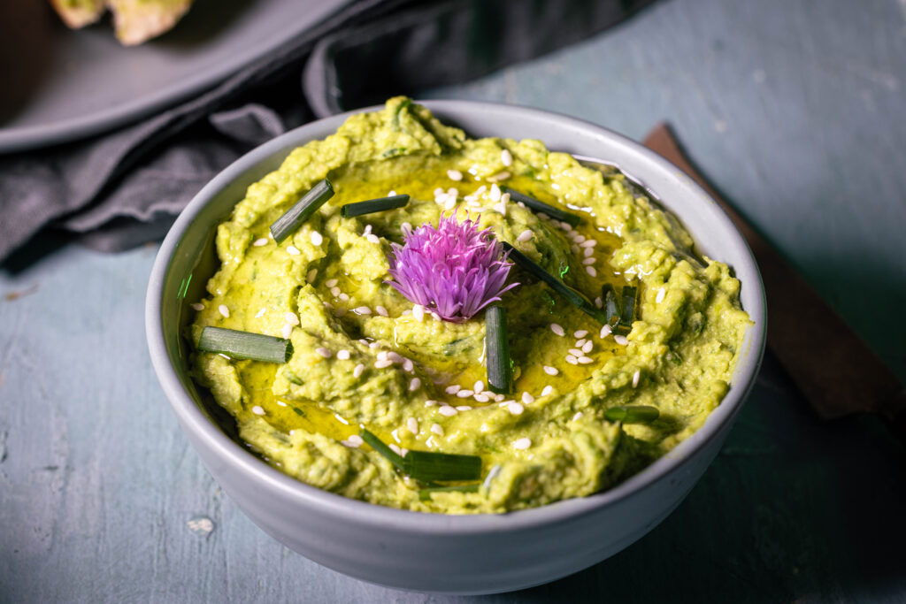 Schnittlauch-Hummus in einer grünen Schale, garniert mit Olivenöl, Schnittlauchröllchen und einer lila Schnittlauchblüte.