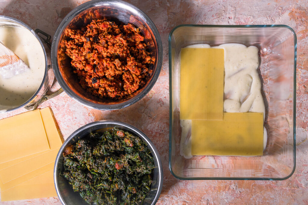 Eine vegane Wildkräuterlasagne wird in einer Auflaufform geschichtet.