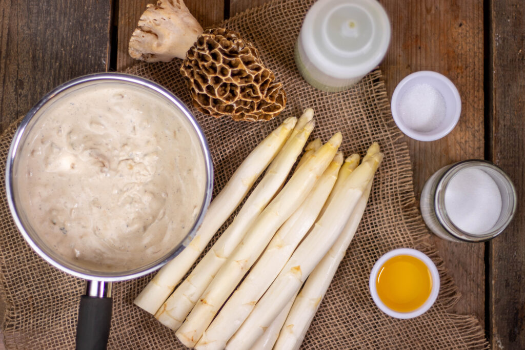 Weißer Spargel, eine Speisemorchel, ein Topf mit Morchelsauce und weitere Zutaten auf einem Holztisch.
