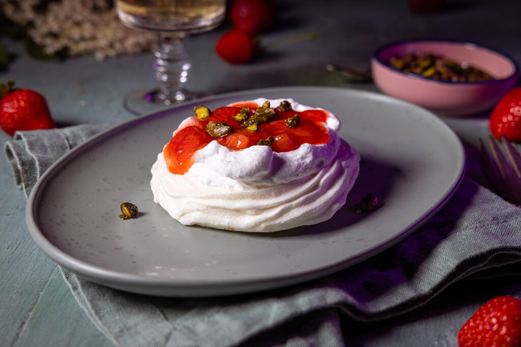 Erdbeer Pavlova auf einem grünen Teller. Im HIntergrund ein Getränke, Erdbeeren und ein Schälchen mit karamellisierten Pistazien.